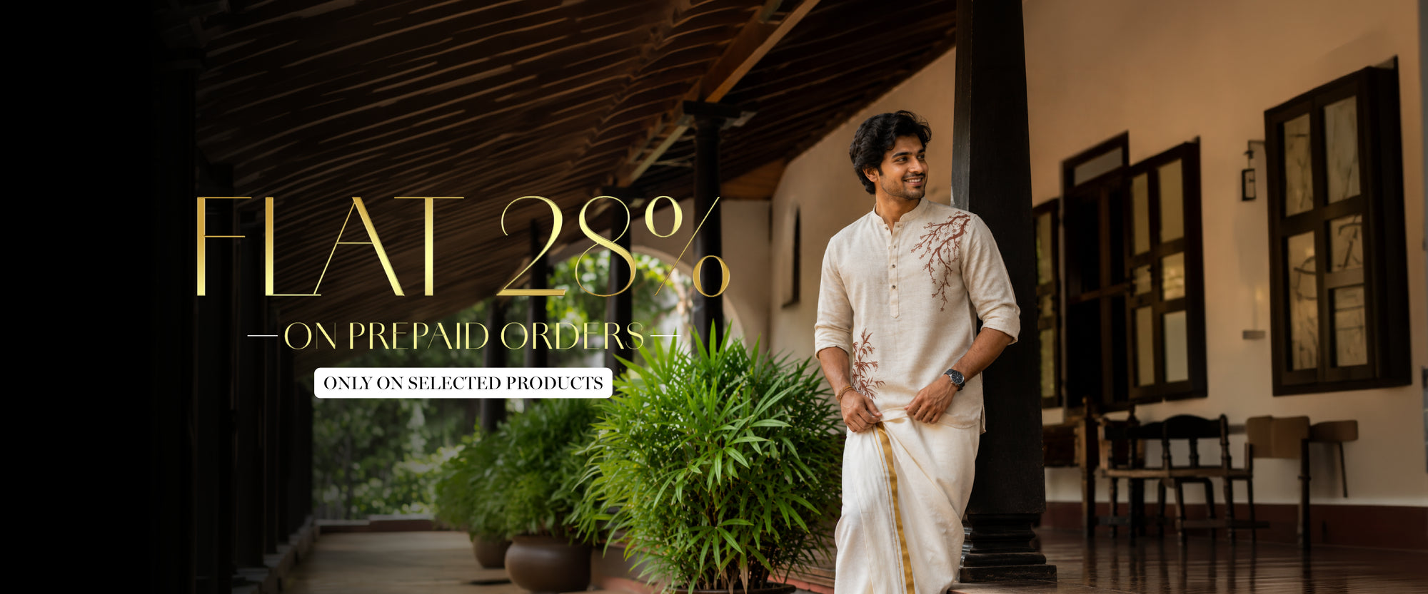 A man in a white embroidered shirt for onam and drape stands on a porch with large potted plants and dark columns. L
